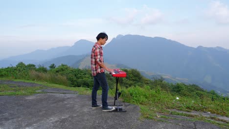 A-stationary-footage-of-a-man-from-his-right,-playing-his-electronic-musical-keyboard-with-a-mountain-view-in-the-background