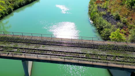 Aerial-view-of-grassy-abandoned-railway-track-on-bridge-over-calm-clean-river