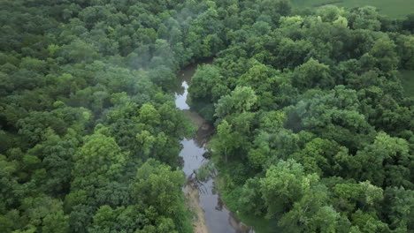 Flyover-above-small-river-winds-through-forest-towards-field-in-rural-Missouri