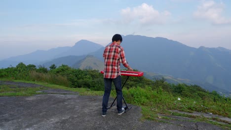 A-stationary-footage-of-a-man-from-his-rear,-playing-his-electronic-musical-keyboard-with-a-mountain-view-in-the-background