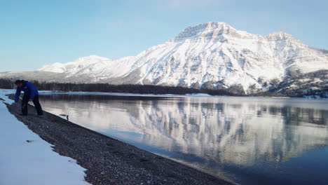 Man-with-blue-jacket-skipping-rocks-on-lake,-snowy-mountain-backdrop