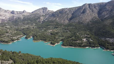 Dramatic-blue-turquoise-lake-surrounded-by-mountainous-terrain-and-forest,-Sunny