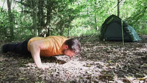 Young-strong-man-doing-push-ups-in-forest-campsite-with-tent-in-background