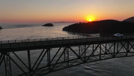 Aerial-shot-of-a-white-truck-driving-over-Deception-Pass-State-Park-at-sunset