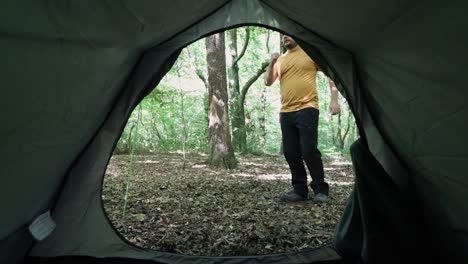Man-stretching-and-preparing-to-go-for-a-job-in-woodland-area,-view-from-inside-tent-on-a-bright-day