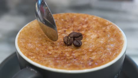 With-the-help-of-a-spoon,-breaks-the-crystalized-hard-brittle-layer-of-caramel-of-a-cup-of-Brulee-coffee-and-decorated-with-coffee-beans