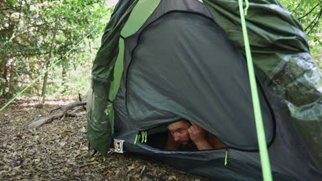 Scared-man-unzip-tent-door-and-carefully-observe-surroundings-in-dense-forest-landscape