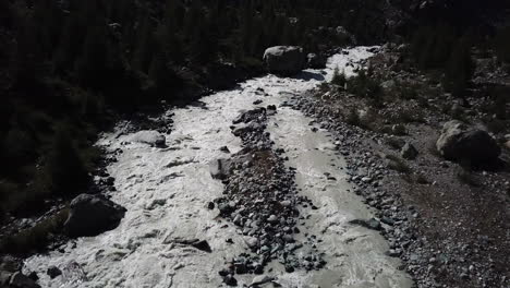 Riverbed-of-a-muddy-river-with-strong-current-and-rocks,-valley-in-the-Swiss-alps,-tilt-up-drone-aerial-view