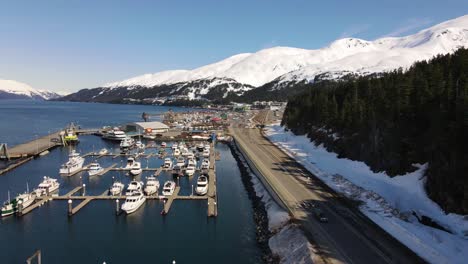 Drone-ascending-over-a-marina-in-Whittier-Alaska-pine-trees-and-snow-capped-mountains-in-the-background