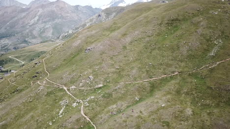 Drone-aerial-view-of-a-grass-field-in-the-Swiss-alps,-hiking-gravel-path,-rocky-summits-for-mountain-enthusiasts,-bike-or-mountain-walks