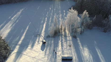 Luftaufnahmen-Von-Oben-Nach-Unten-Von-Einer-Verschneiten-Winterlandschaft-Mit-Holzhüttenbeleuchtung-Durch-Sonnenlicht-Am-Morgen---Eine-Gruppe-Von-Menschen,-Die-Auf-Einem-Verschneiten-Ländlichen-Feld-Spazieren