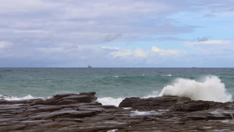 Waves-crashing-onto-the-rocks-whilst-a-container-ship-floats-on-the-horizon-near-Sydney,-Australia