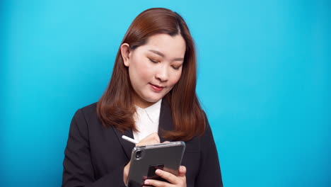 Schöne-Asiatische-Geschäftsfrau-In-Einem-Schwarzen-Anzug,-Der-Ein-Digitales-Tablet-Für-Die-Planung-Von-Online-arbeiten-Mit-Drahtloser-Technologie-Auf-Blauem-Hintergrund-Verwendet