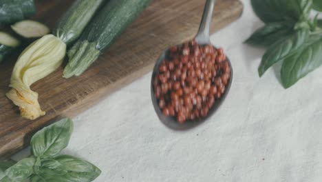 Shot-from-above-of-a-table-on-which-a-silver-spoon-full-of-red-pepper-is-placed,-from-the-overhang-the-grains-fall-all-around,-in-the-scene-there-are-also-courgettes-and-basil-leaves