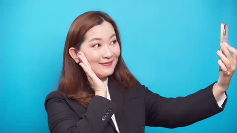 Junge-Glückliche-Asiatische-Geschäftsfrau,-Die-Ein-Smartphone-Verwendet,-Um-Sich-Lächelnd-In-Einem-Schwarzen-Anzug-Auf-Blauem-Hintergrund-Winkend-Zu-Machen