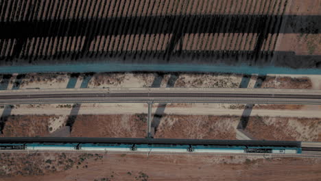 Top-down-shot-of-2-trains-crossing-next-to-each-other-on-a-massive-railway-bridge-over-fields-of-vineyards