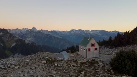 Lone-Cabin-Hut-at-Sunset-on-Mount-Brew-Peak-Surrounded-by-Rocky-Landscape-with-Trees-in-Canada-BC---Retreating-Aerial-Drone-Shot