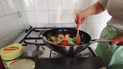 woman-stirs-wooden-spoon-in-frying-pan-full-with-vegetables-broccoli-and-peppers