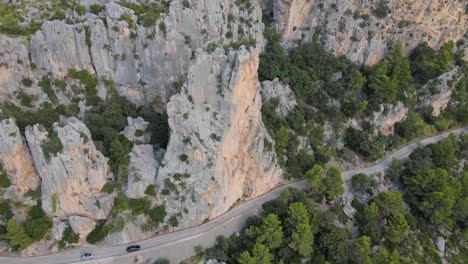 Birds-eye-view-of-cars-driving-down-a-mountain-road-with-spiky-peaks-towering-out-beside-them-at-Sa-Calobra,-Mallorca,-Spain