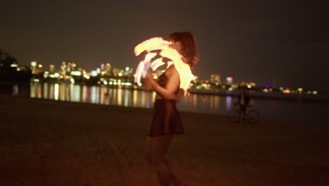 Woman-Twirling-Fire-Fans-On-Lakeshore-Beach-Against-Toronto-Skyline,-Exterior-Night-Medium-Shot