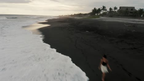 Schwarzer-Sandstrand-Mit-Schäumenden-Wellen,-Die-Bei-Sonnenuntergang-In-El-Paredon,-Guatemala,-An-Land-Spritzen---Drohnenaufnahme
