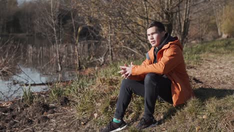 Young-man,-with-an-orange-jacket,-sits-near-the-lake-and-contemplates-the-panorama