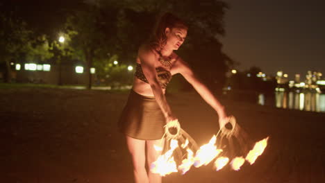 Woman-Dancing-With-Fire-Fans-on-Lakeside-Beach-Against-Toronto-Skyline,-Exterior-Night-Medium-Shot