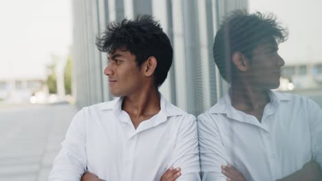 Indian-young-guy-crossing-his-arms,-leaning-against-a-glass-wall-on-the-street