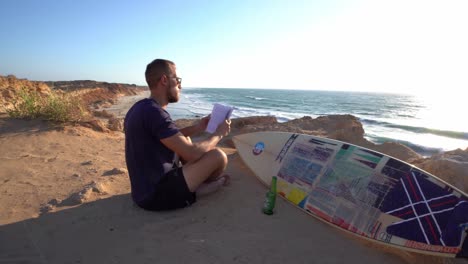 Rotating-cinematic-shot-of-a-person-reading-a-book-sitting-near-a-surfboard-and-beer-bottle-at-a-beach-in-Hadera,-Israel