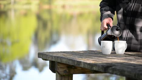 Man-in-checked-flannel-pouring-coffee-from-french-press-into-two-mugs,-outdoors