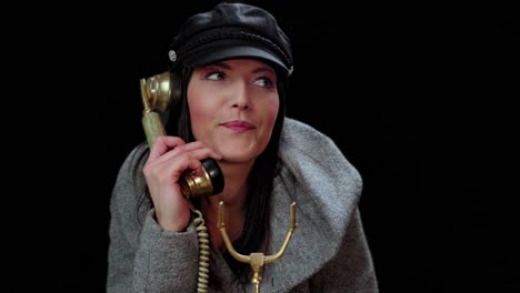 Close-up-shot-of-a-woman-with-black-cap-and-grey-dress-talking-on-old-vintage-phone-smiling-with-black-background