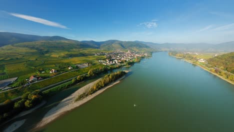 FPV-ascending-over-the-glistening-river-Danube,-smoothly-approaching-beautiful-market-town-Spitz,-Wachau,-Austria