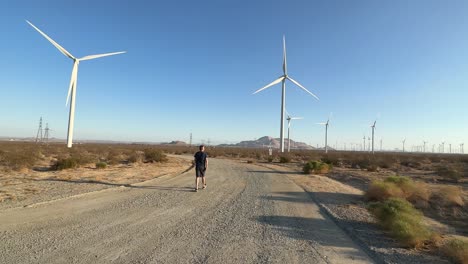 Man-Walking-on-Dirt-Road-in-Windmill-Park,-Spinning-Blades-of-Wind-Turbines-on-Sunny-Day,-Static-Shot