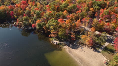 Aerial-drone-shot-flying-above-a-campsite-area-surrounded-by-colorful-trees-of-fall-next-to-mirrored-lake-and-people-standing-and-observing-the-beauty,-Canada,10-Oct,-2022