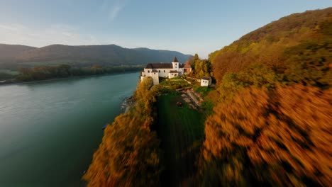 FPV-soaring-above-stunning-autumn-woods-towards-painterly-monastery-Schönbühel,-diving-down-in-a-dynamic-rotation-next-to-it