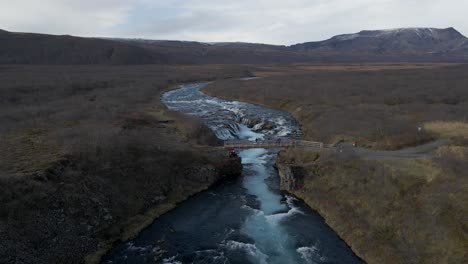 Gente-En-El-Puente-De-Madera-Sobre-La-Cascada-De-Bruararfoss-Y-El-Paisaje-Circundante,-Islandia