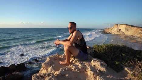 Ein-Mann-In-Sportbekleidung-Und-Schwarzer-Sonnenbrille-Sitzt-Am-Felsigen-Strand-Und-Blickt-Auf-Den-Blauen-Ozean-In-Haderah,-Israel