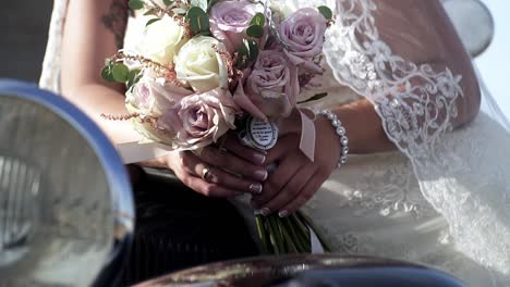 Slow-motion-handheld-shot-of-the-bride-dressed-in-white-wedding-dress-and-jewelry-on-her-hand-leaning-against-a-black-vintage-car-with-a-bouquet-of-roses-in-her-hand-on-wedding-day