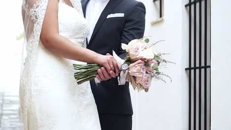 Static-slow-motion-handheld-shot-of-a-wedding-couple-while-the-groom-holds-the-hand-of-his-bride-with-a-bouquet-of-roses-in-his-hand-on-a-bright-sunny-day