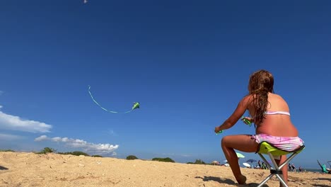 Red-haired-little-girl-sitting-on-beach-chair-enjoys-flying-a-kite-on-hot-summer-day