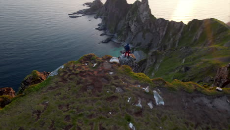 Stunning-orbiting-aerial-view-of-a-person-walking-on-the-edge-of-a-cliff-looking-down-on-the-rocky-coast-with-green-grass-and-enjoying-the-beautiful-sunset-and-scenic-views
