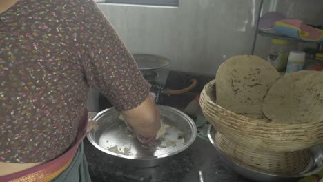 Over-the-shoulder-shot-of-a-senior-aged-woman-preparing-traditional-bhakri-dough-in-Indian-kitchen-using-cooking-gas-stove