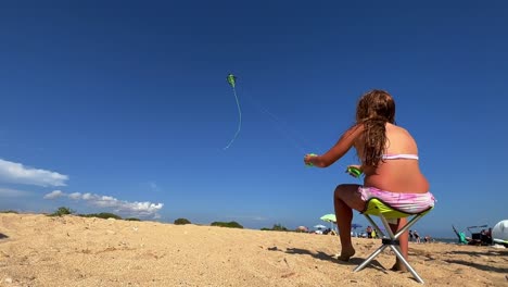 Red-haired-little-girl-sitting-on-small-beach-chair-enjoys-flying-a-double-strand-kite-on-hot-summer-day