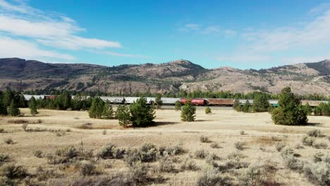 dolly-forward-drone-shot-flying-towards-a-railroad-station-in-a-desert-environment-on-a-sunny-day-with-mountains-and-trees-in-the-background-and-a-bush-and-grassland-in-the-foreground