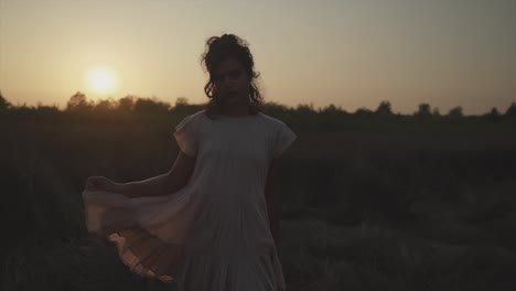 Slow-motion-handheld-shot-of-a-young-indian-woman-in-a-dress-standing-on-a-field-during-a-beautiful-sunset