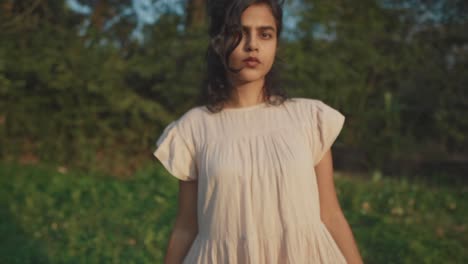 Slow-motion-backward-dolly-shot-of-a-young-charming-indian-woman-in-a-dress-looking-at-the-camera-with-a-serious-look-while-she-is-in-nature