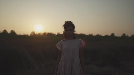 Slow-motion-handheld-shot-during-sunset-of-a-young-pretty-woman-in-a-dress-standing-on-a-field-looking-at-the-camera-in-the-background-the-sun-and-trees