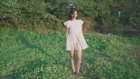 Slow-motion-static-shot-of-a-young-pretty-indian-woman-dressed-in-a-pink-dress-standing-in-the-garden-in-the-grass-between-trees-in-the-background-looking-at-the-camera