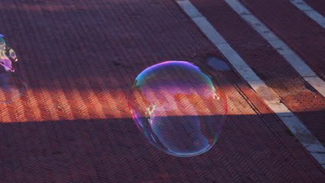 Soap-Ball-Bubbles-Floating-in-Air,-Dragged-by-Breeze-at-Low-Altitude,-Sinking-Down-Towards-Ground,-Colorful-Transparent-Shape-in-Morning-Sunlight