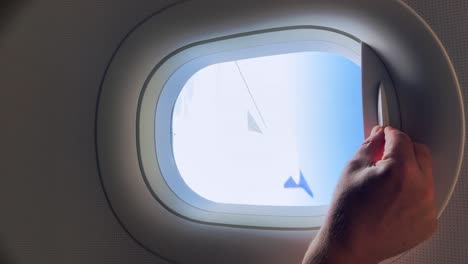 Passenger-hand-opening-aircraft-window-blind-from-which-airplane-wing-and-clouds-can-be-seen